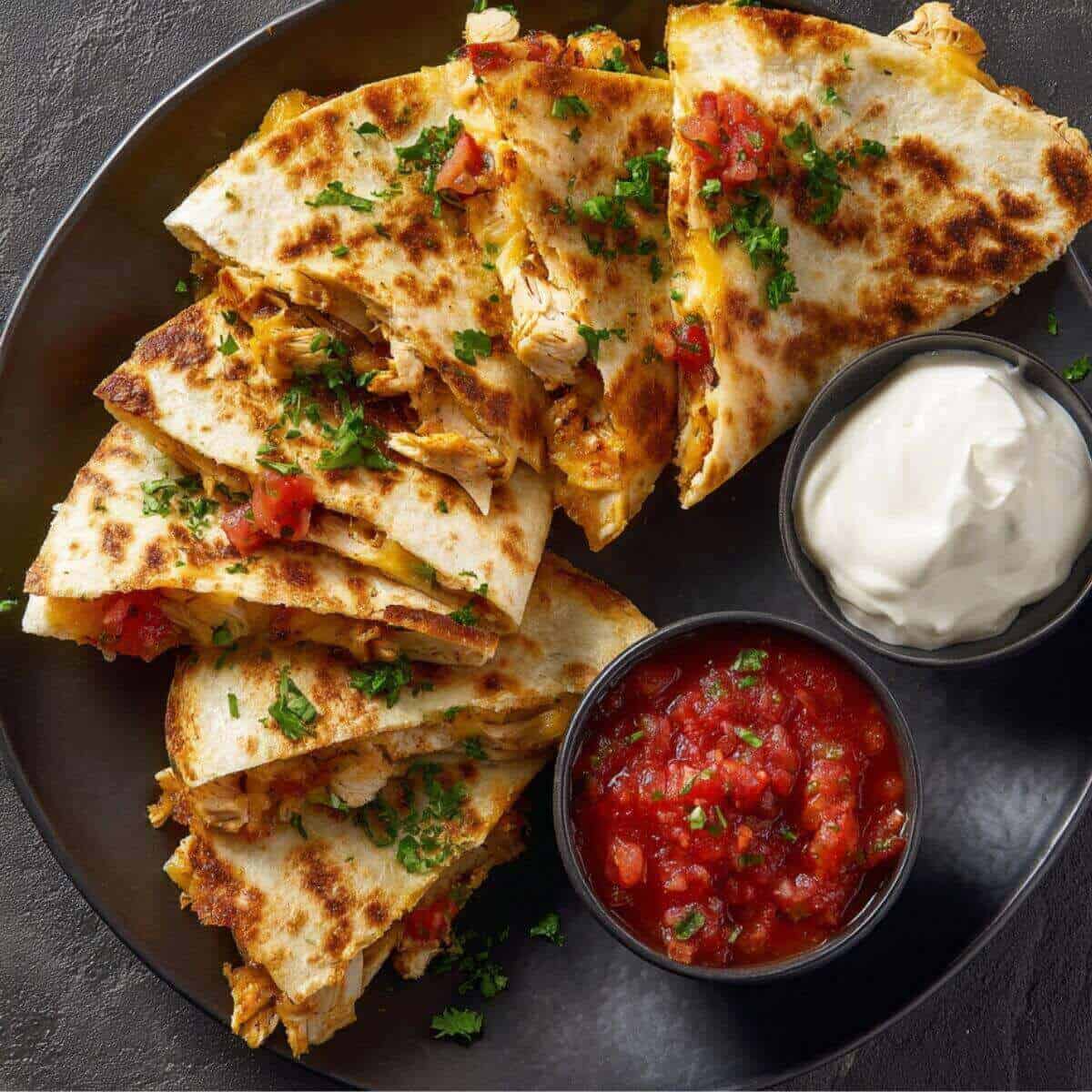 Platter of crispy 30 Minute Chicken Quesadillas garnished with herbs, served with salsa and sour cream.